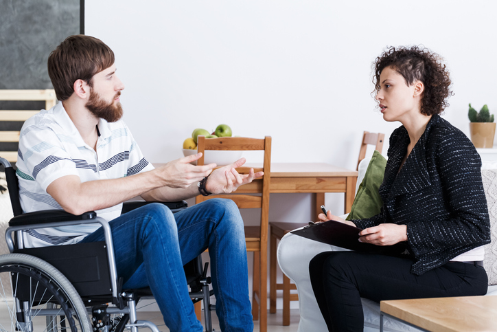Young bearded man in wheelchair talking with psychotherapist at home.
