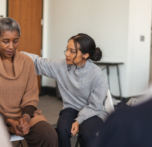 A multi-ethnic group of adults are attending a group therapy session. The attendees are seated in a circle. A senior black woman is sharing her struggles with the group. A mixed-race young woman rests her arm on the woman's back, expressing comfort and support.