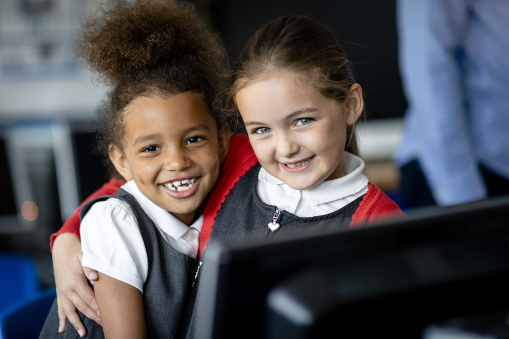 A close up front-view of two best friends sitting in computer class with their arms affectionately over each other's shoulders and smiling as they look towards the camera.